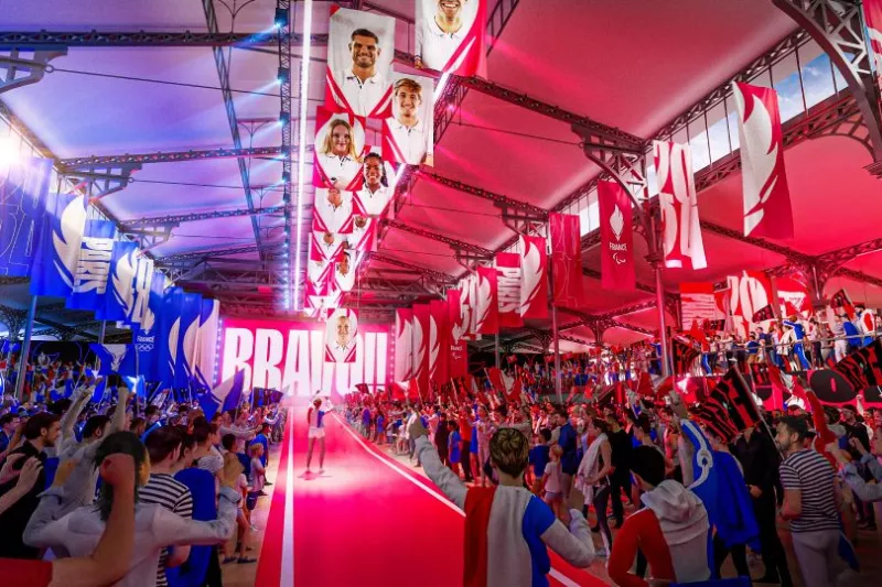 alt="Image de synthèse de l'intérieur de la Grande Halle de la Villette en mode célébration des athlètes avec un couloir central et les supporters de chaque côté. La Halle est au aux couleurs du Club France en Bleu, Rouge. Il y a des drapeaux à l'effigie des athlètes Français suspendus au plafond."